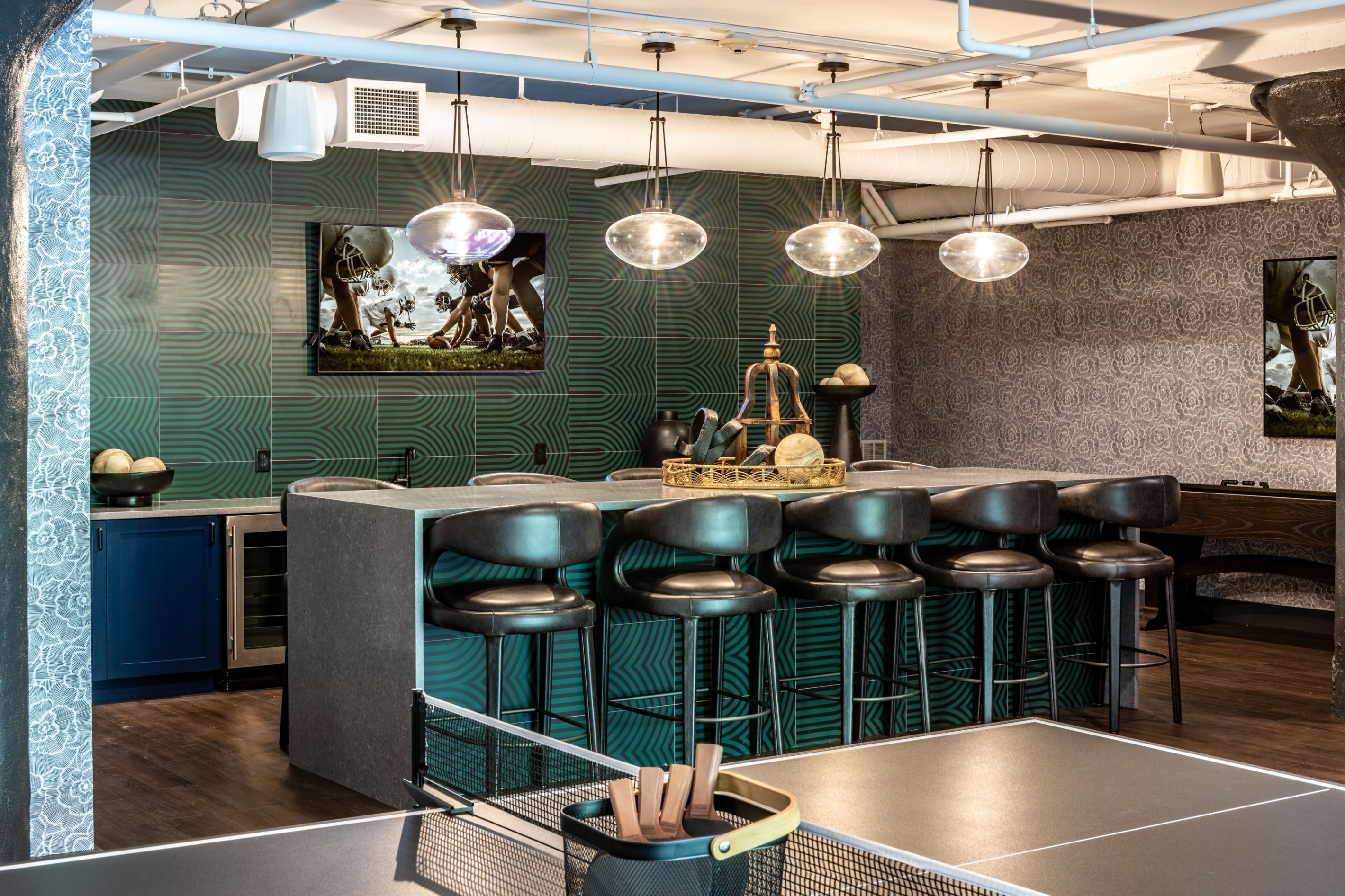 Modern bar area with green tiles, black stools, and overhead pendant lights.