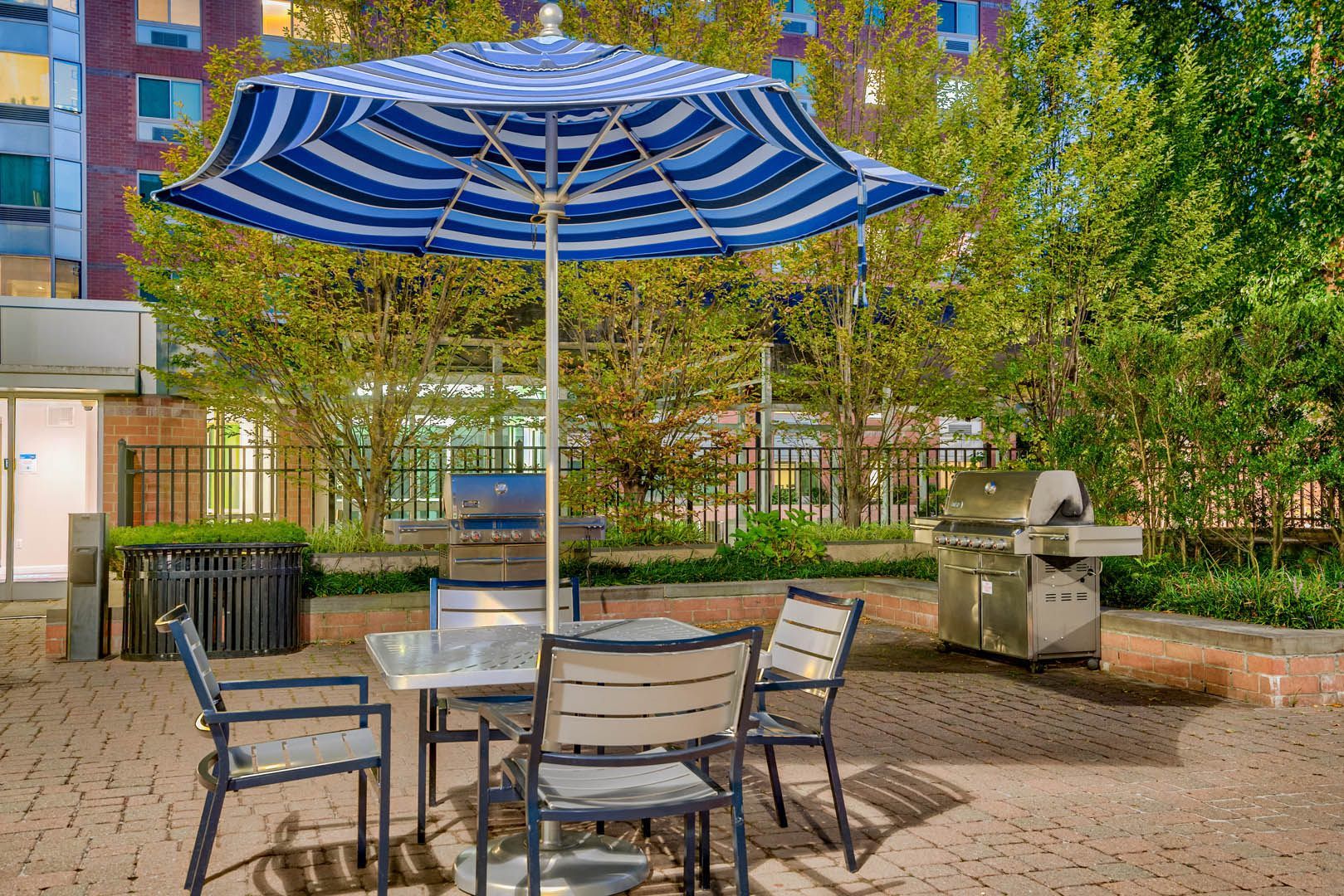 A patio with a table and chairs under a striped umbrella, surrounded by greenery.