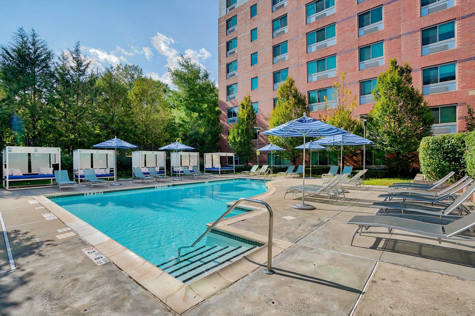 Hotel pool area with umbrellas, lounge chairs, and greenery in the background.