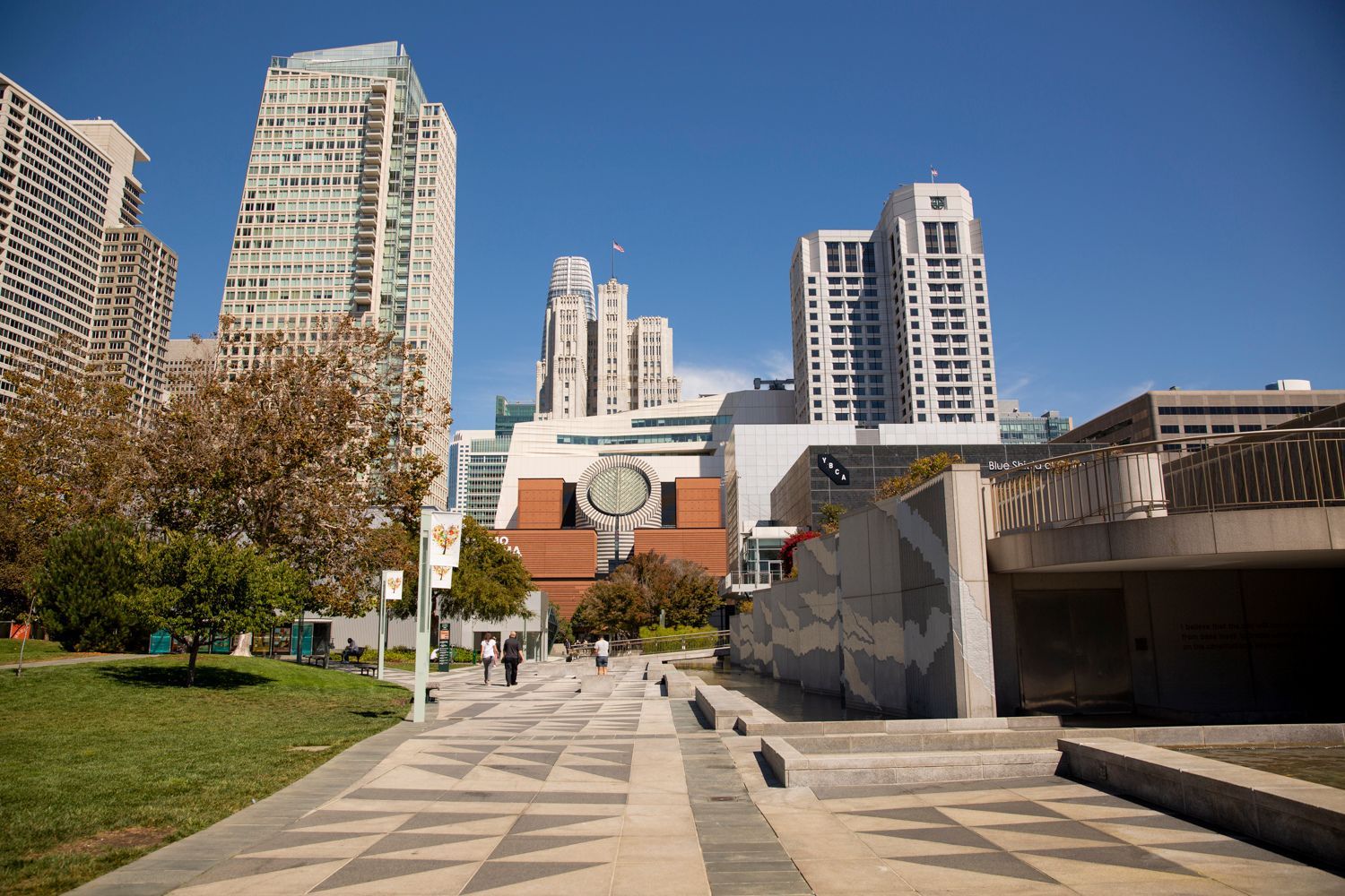 City skyline with modern buildings against a clear blue sky and a paved walkway.