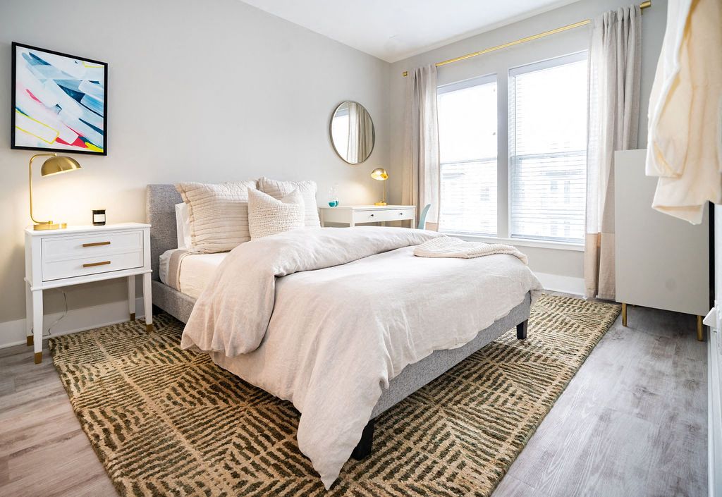 Bright bedroom with a queen bed, nightstand, and large window, accented by a rug.