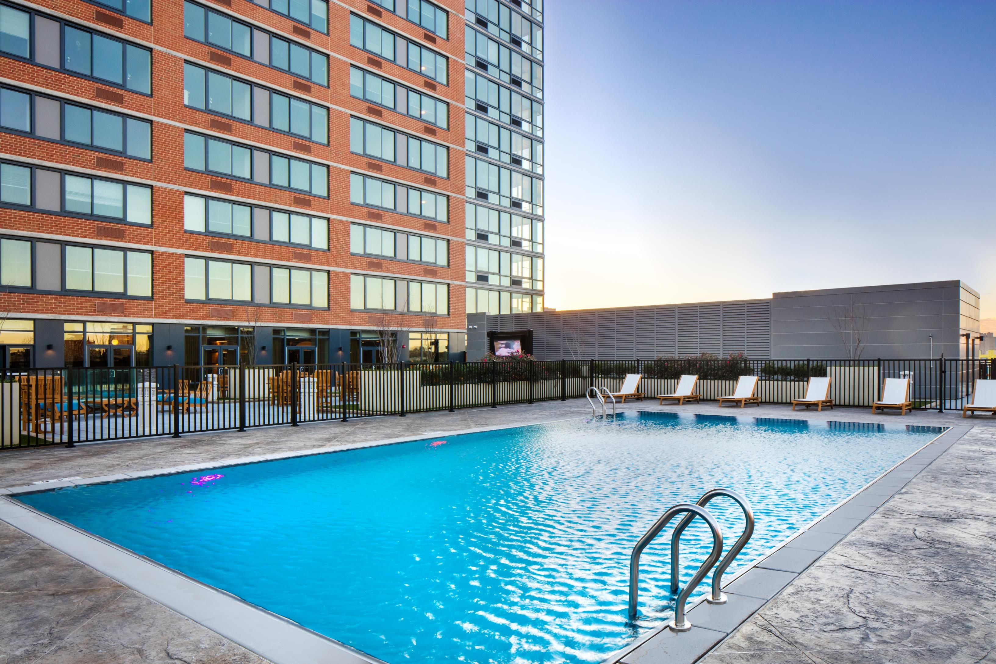 Outdoor pool with lounge chairs and metal fence beside red brick mid-rise at sunset
