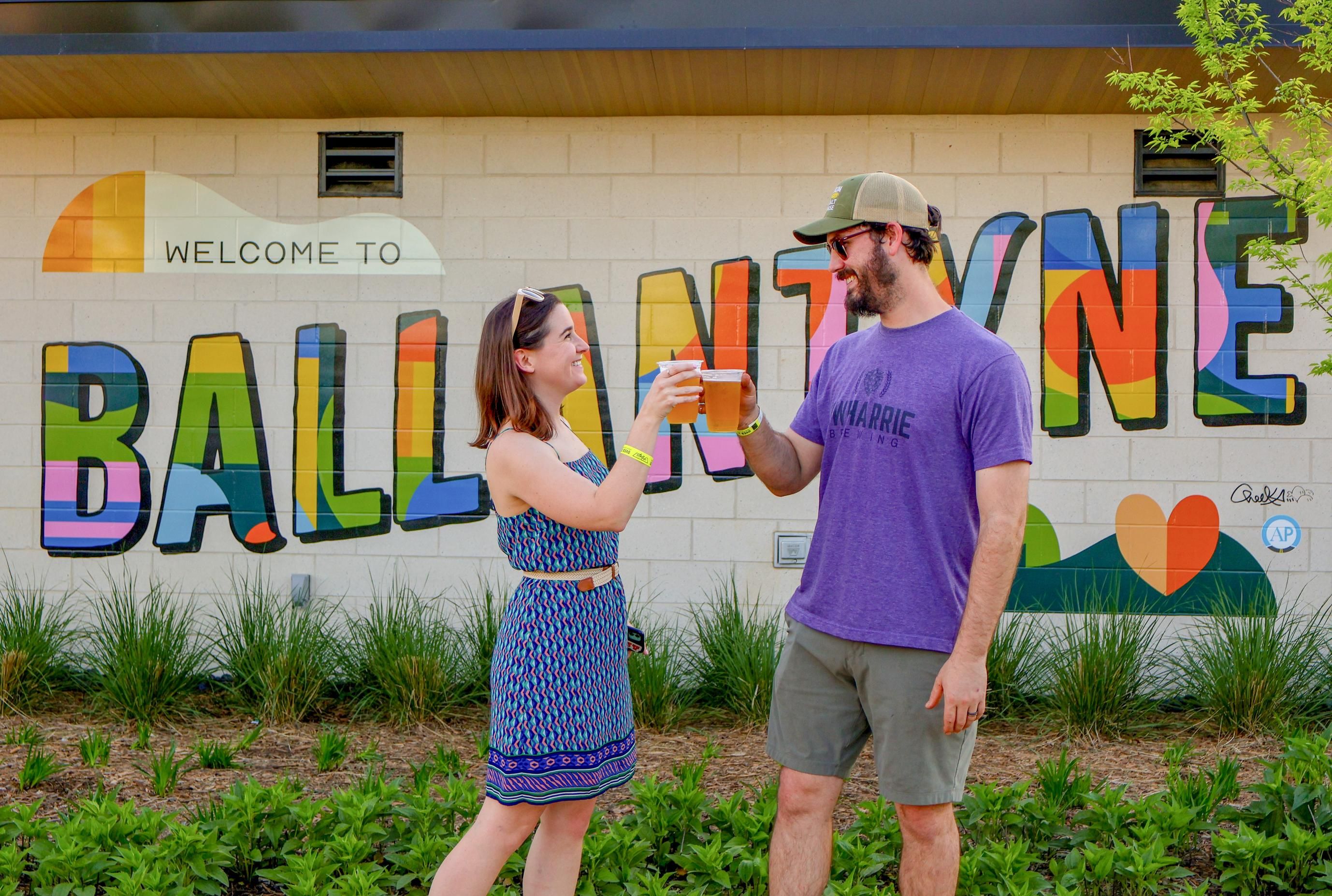 A couple toasting drinks in front of a colorful mural that says "Ballantyne."