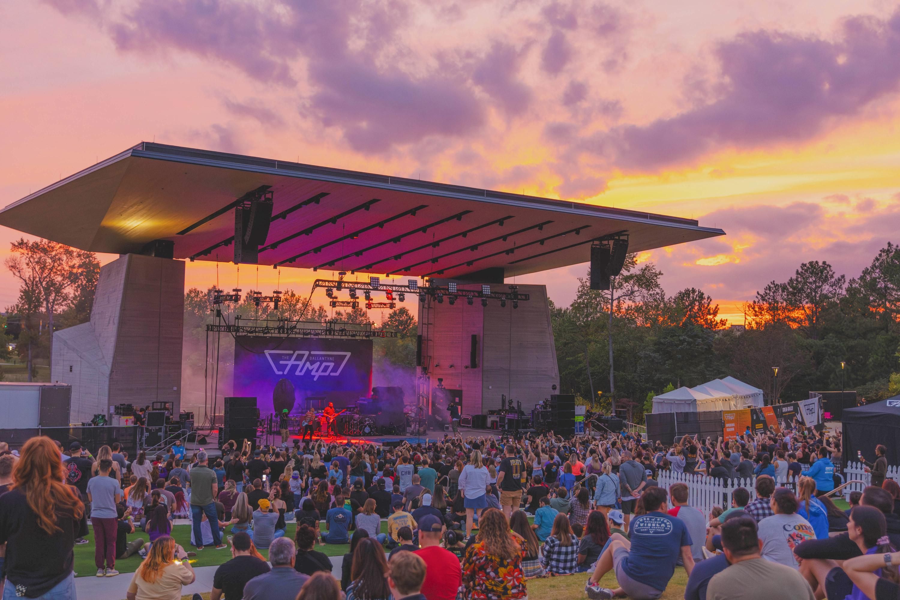 Outdoor concert at sunset with a large crowd and a stage under a modern canopy.