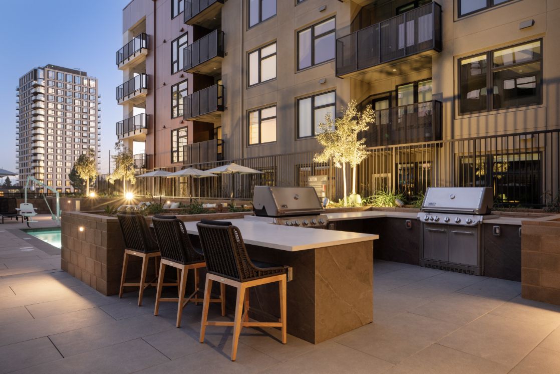 Modern outdoor kitchen and seating area at dusk, with an apartment building in the background.