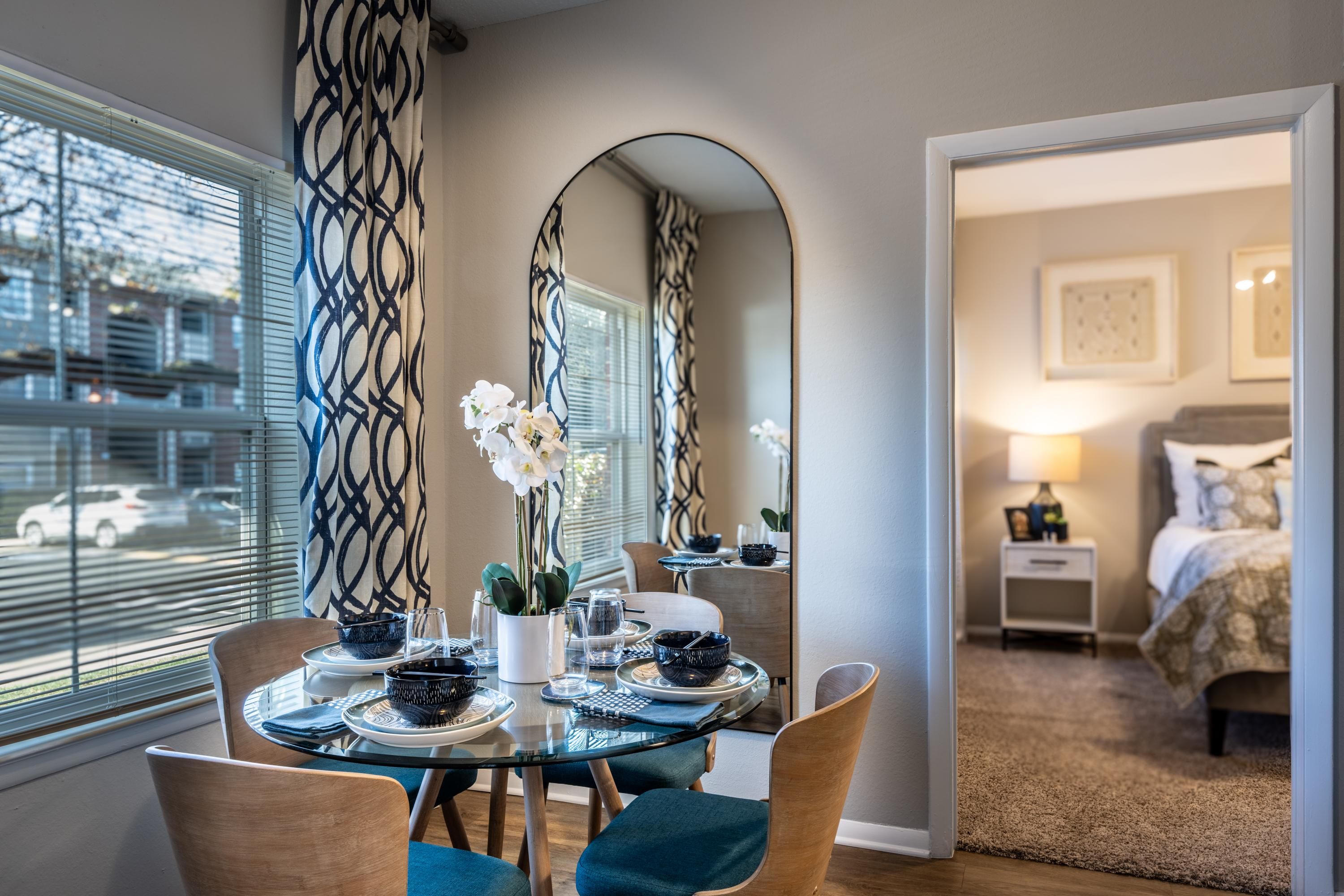 Bright dining area with a mirror, glass table, and a glimpse of a bedroom.