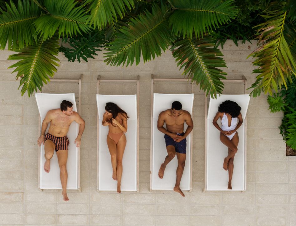 Four people relax on lounge chairs under palm leaves.