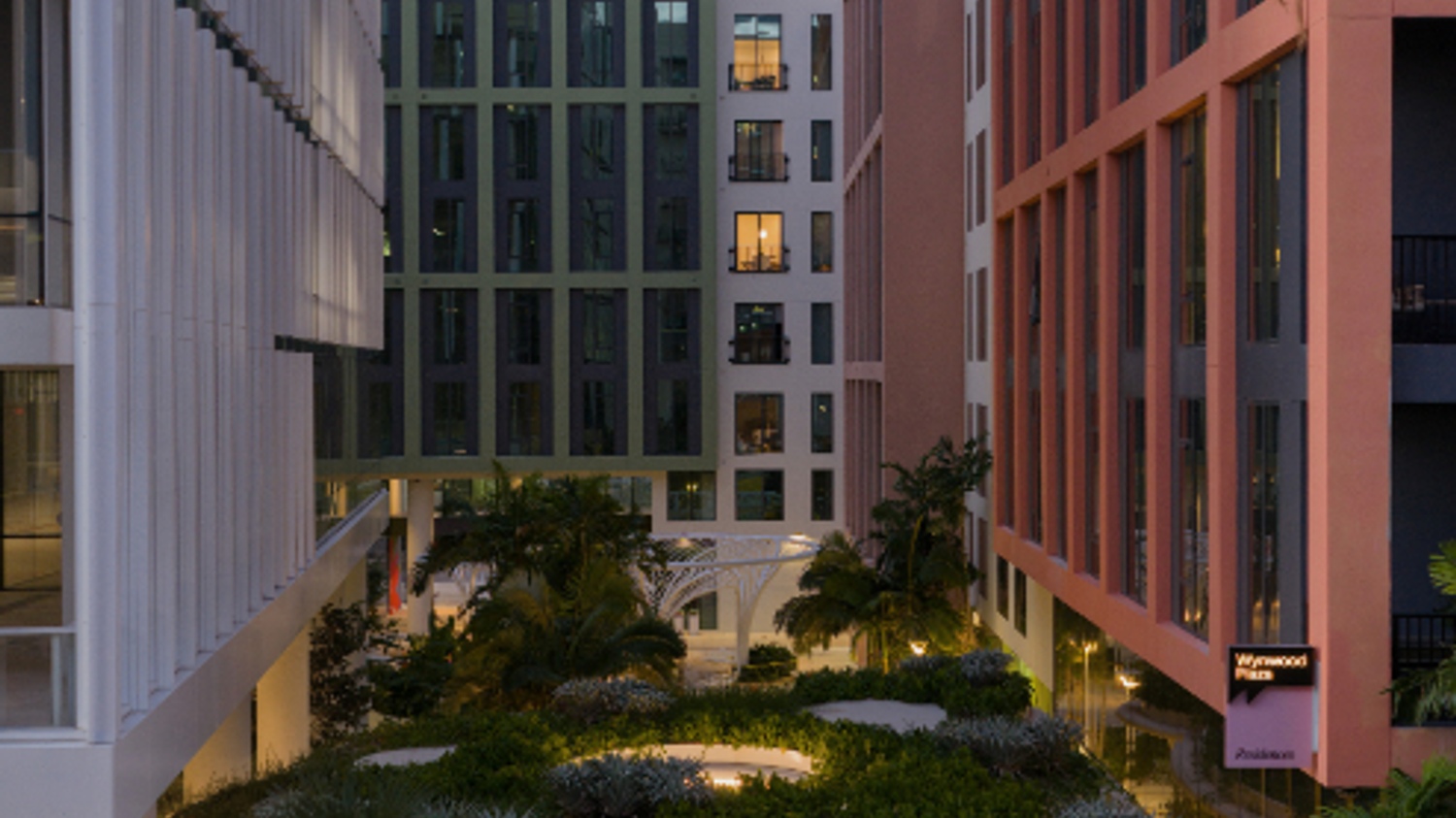 Urban courtyard surrounded by modern buildings with greenery and evening lighting.