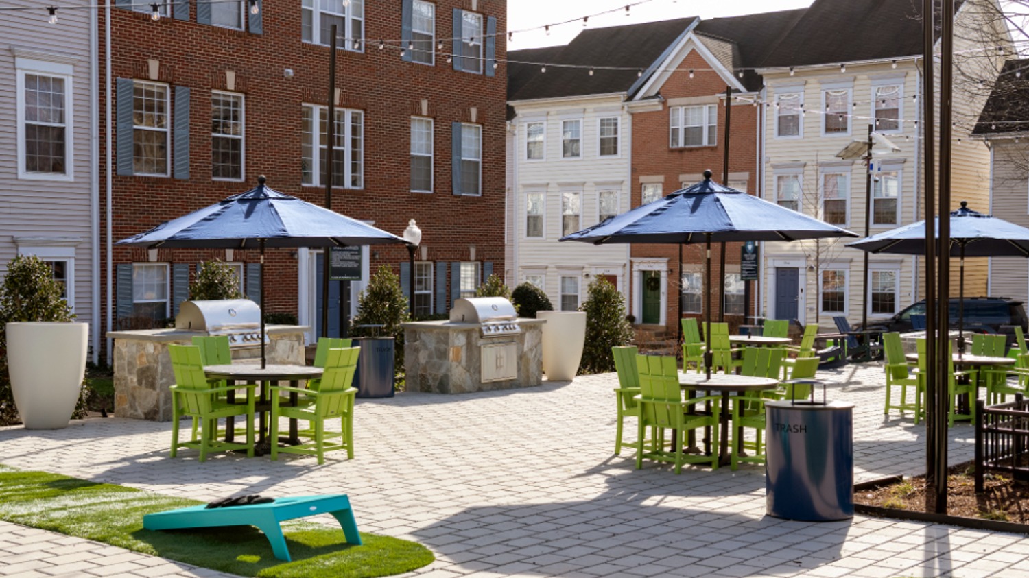 Outdoor lounge area with green furniture, grills, and umbrellas near apartment buildings.