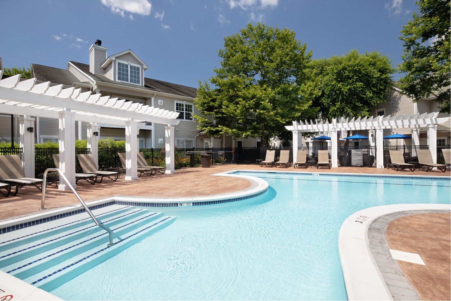 A serene poolside with loungers, surrounded by trees and pergolas under a clear blue sky.