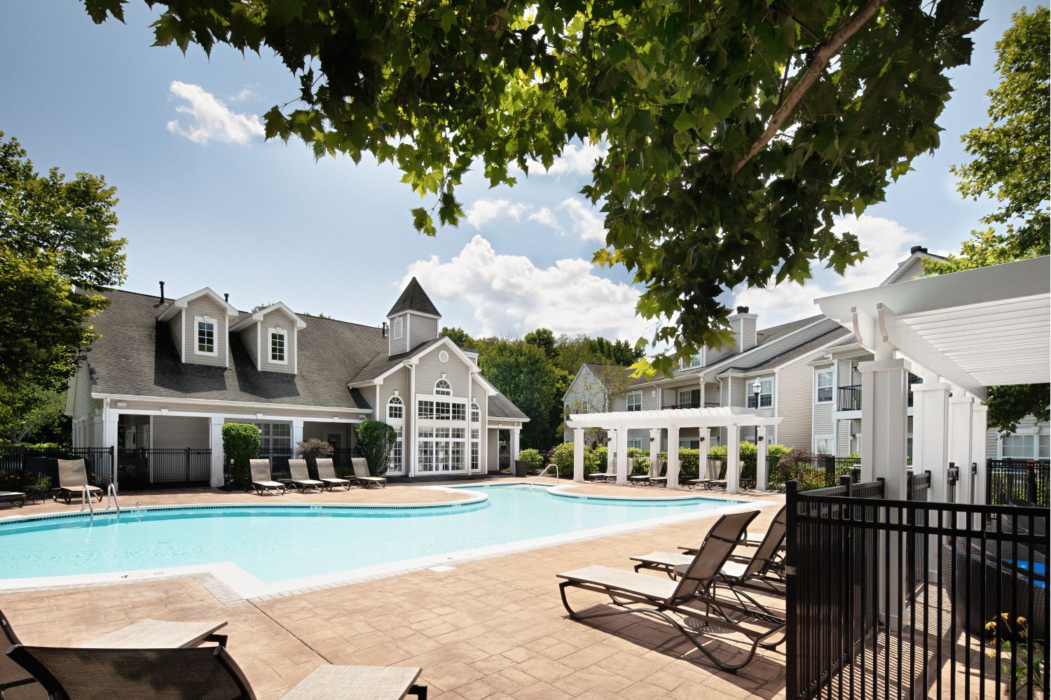 Outdoor pool area with lounge chairs and trees, surrounded by buildings under a blue sky.