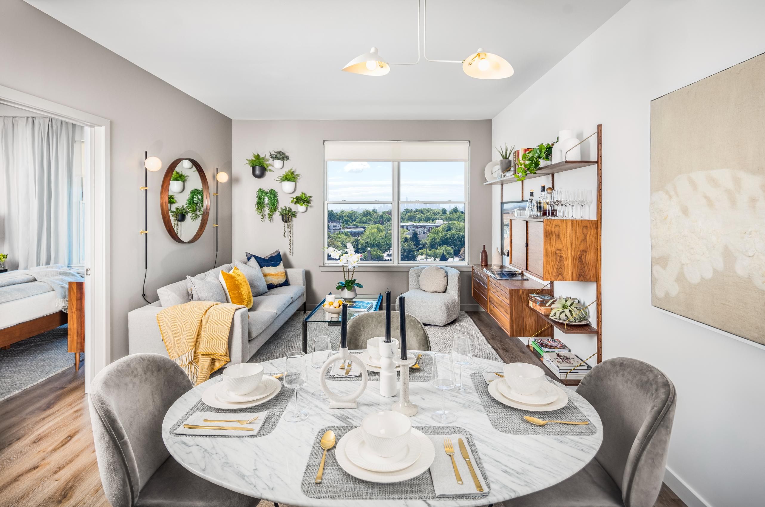 Modern living room with dining area, neutral tones, and large window showing green landscape.