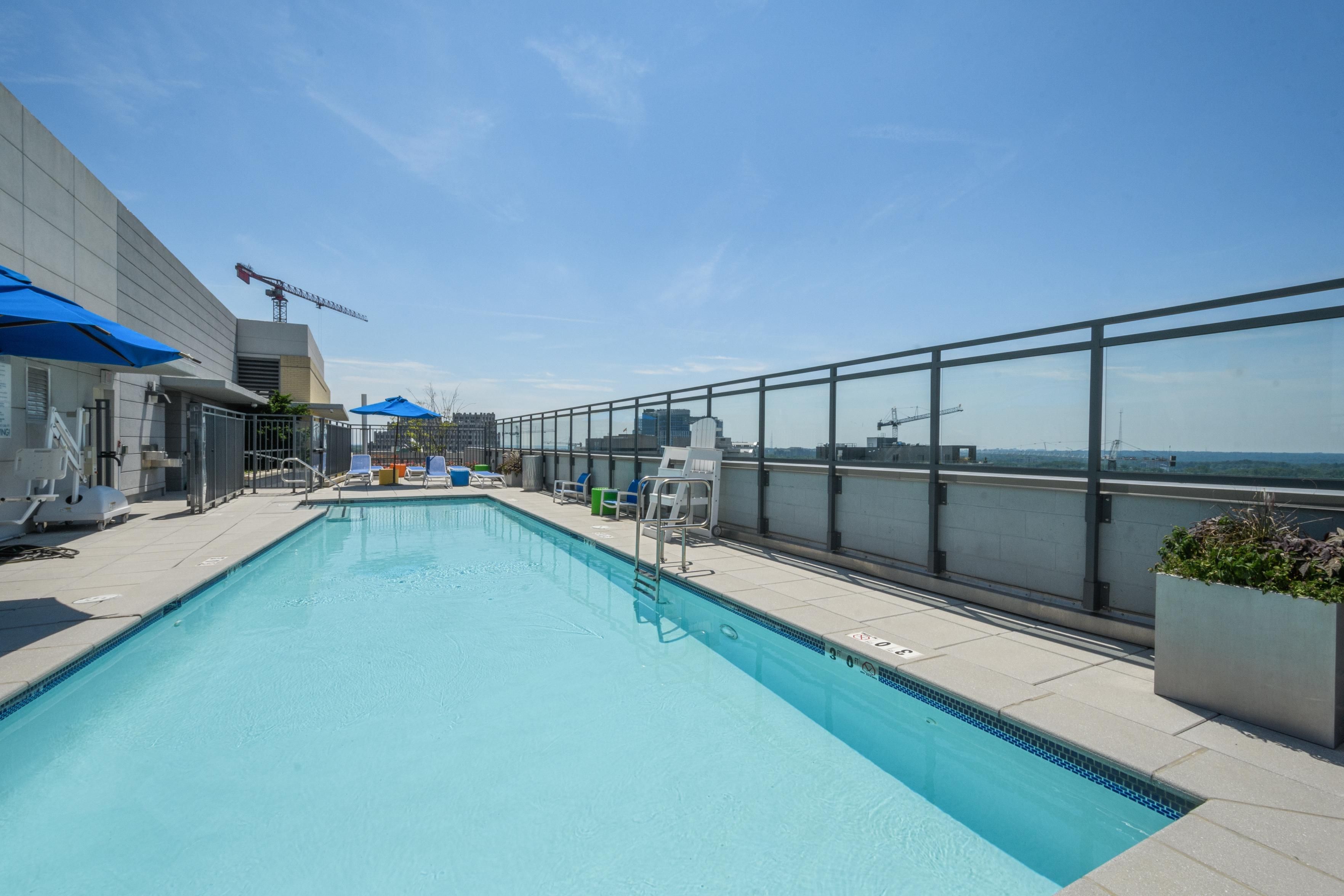 Rooftop pool with lounge chairs under blue sky and a city skyline view.
