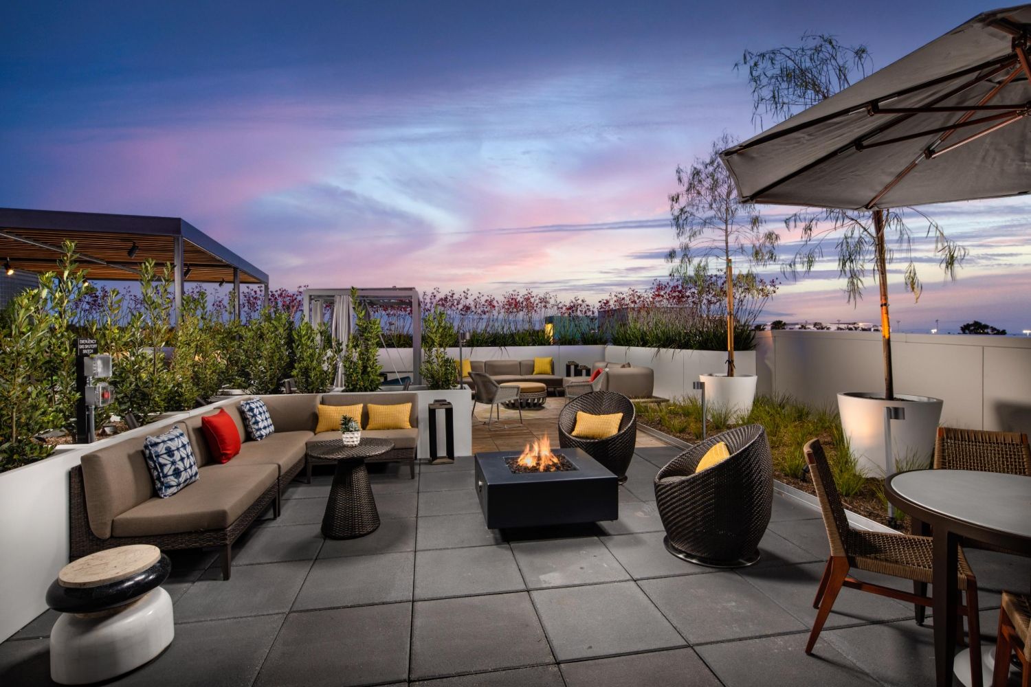 Rooftop lounge with seating, fire pit, and colorful cushions under a sunset sky.