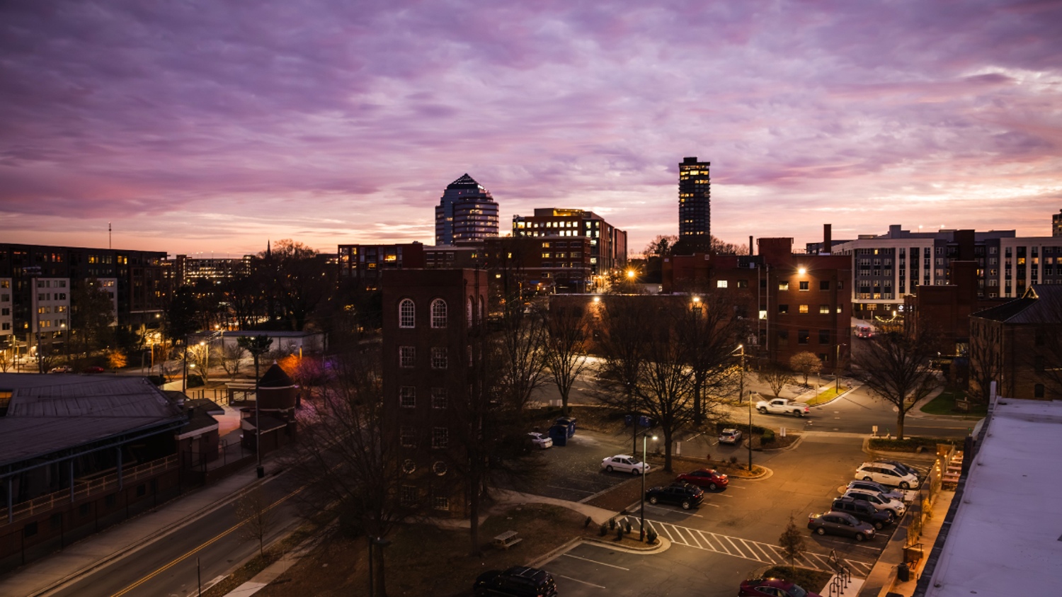 Urban skyline at dusk with purple clouds and warm city lights.