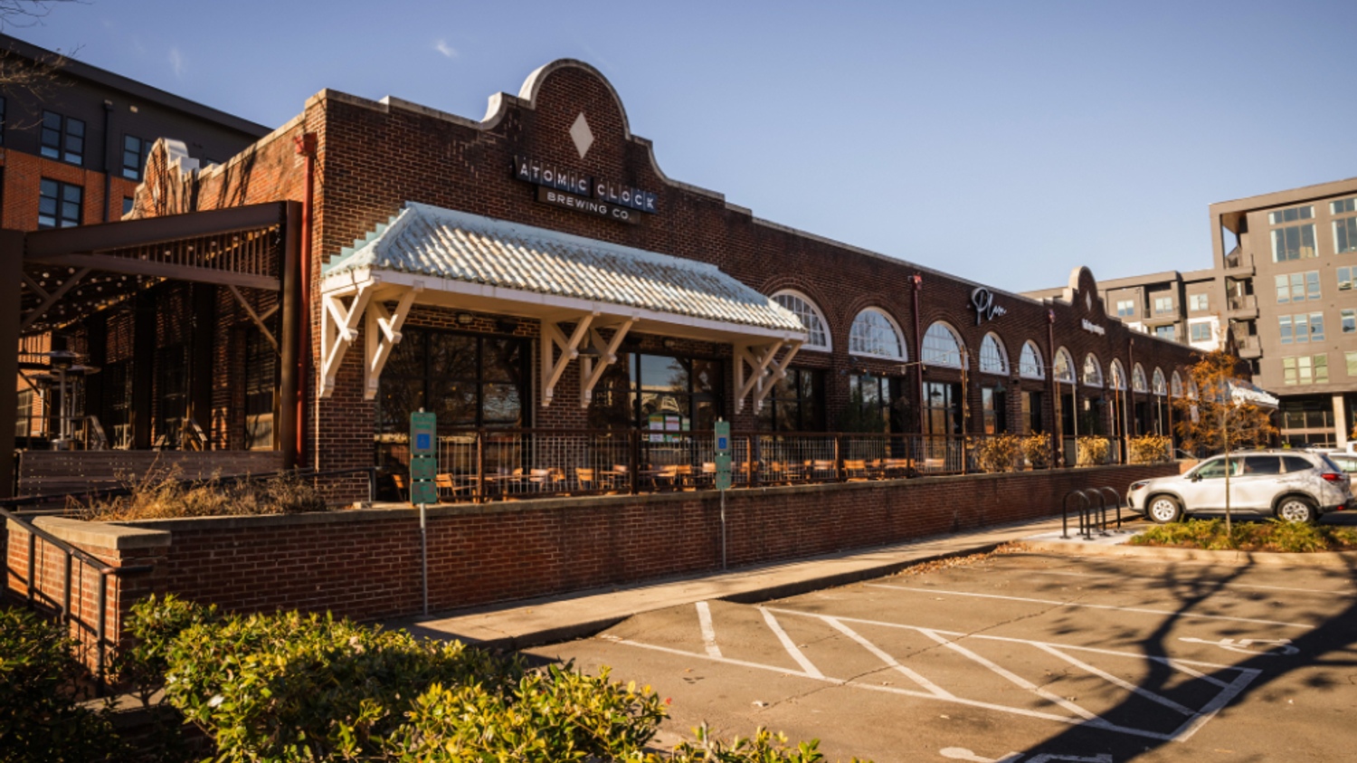 Brick building with large windows and a metal roof, surrounded by a parking lot and greenery.