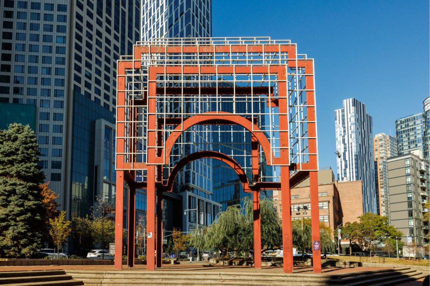 Modern red architectural structure surrounded by city buildings and trees.
