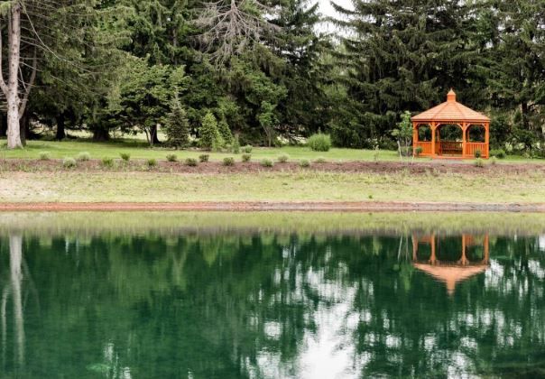 A gazebo reflects on a calm lake surrounded by trees.
