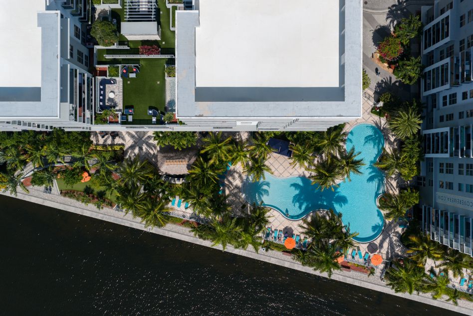 Aerial view of a pool surrounded by palm trees and buildings near a waterway.