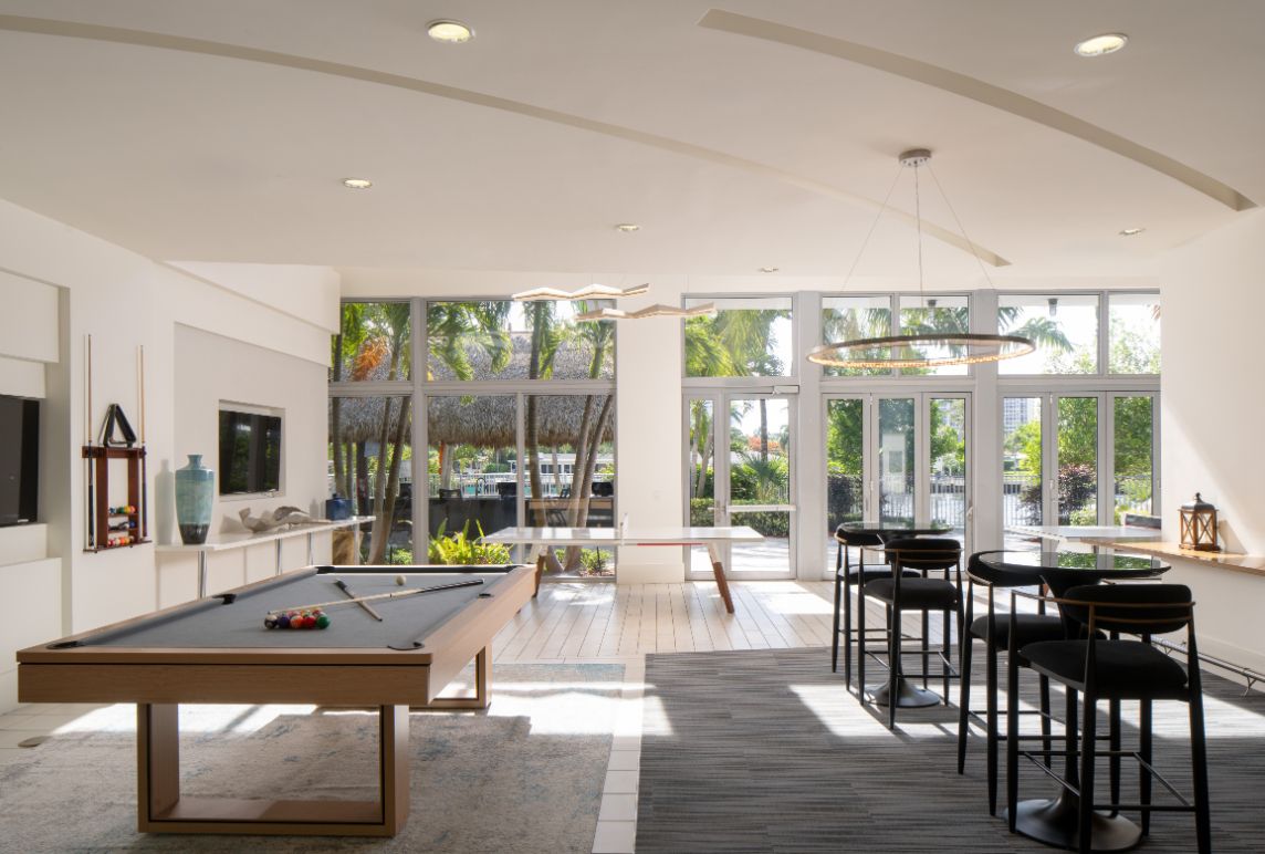 Bright interior with a pool table, bar stools, and large windows overlooking greenery.