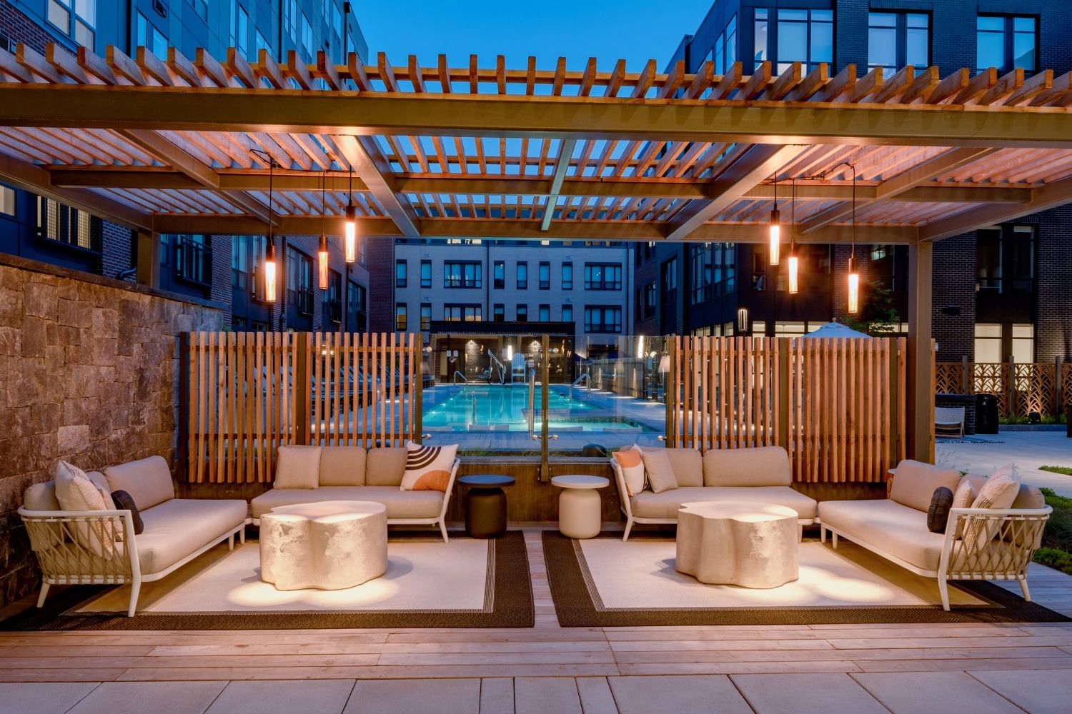 Modern outdoor lounge area with sofas and tables under a pergola by a pool at dusk.