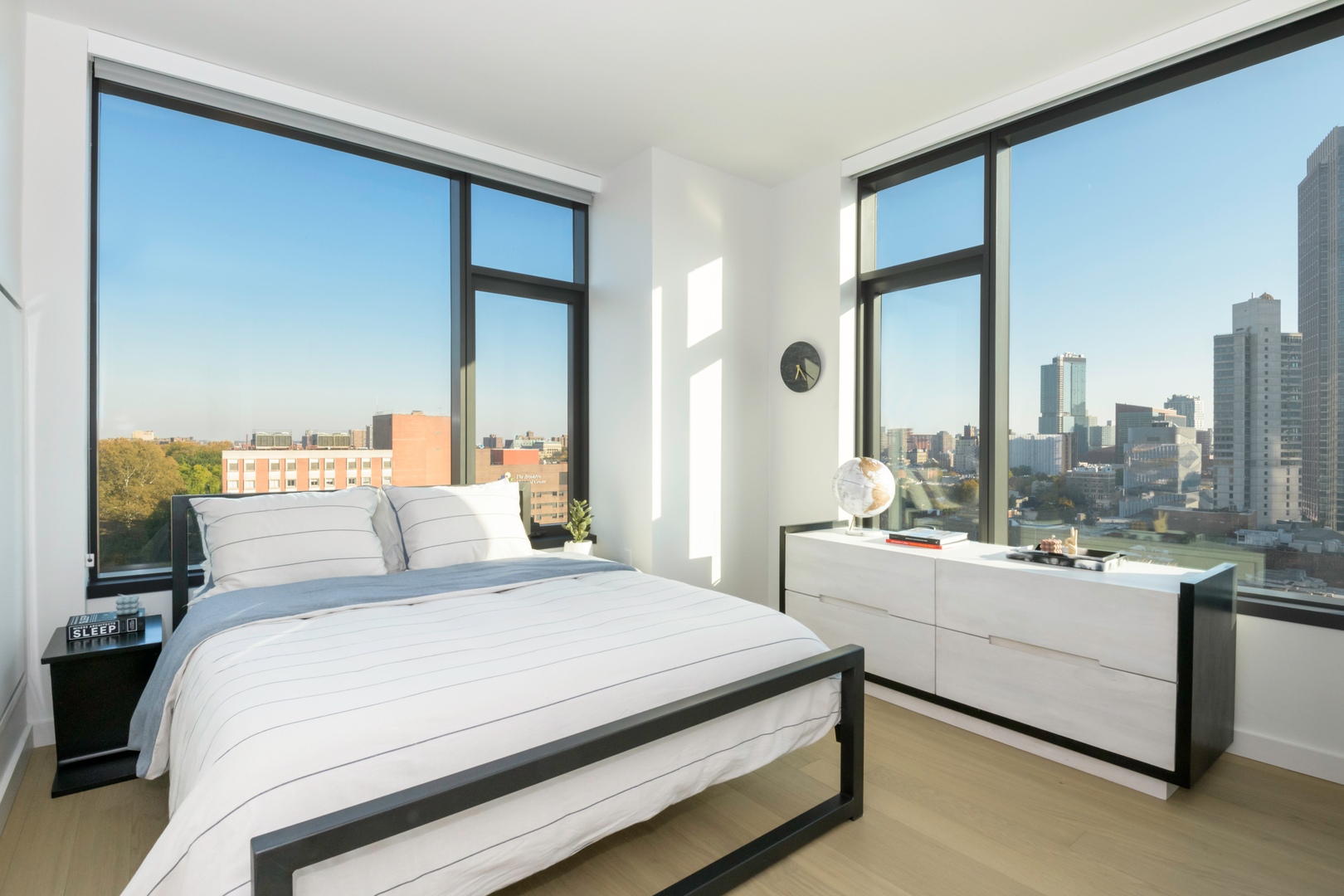 Modern bedroom with a bed, dresser, and large windows showing a city skyline.