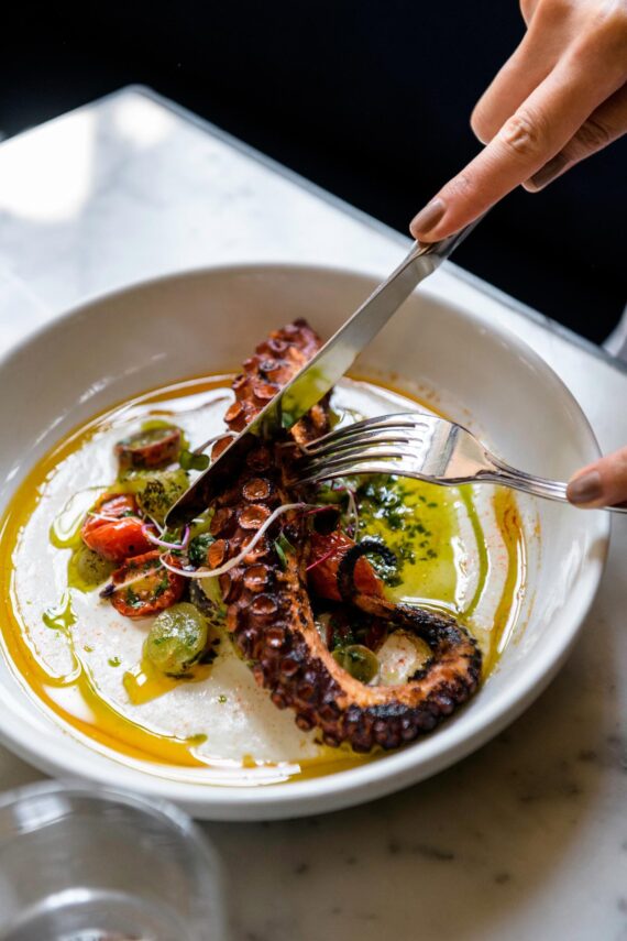 A person cuts into a plate of grilled octopus with vegetables and sauce.