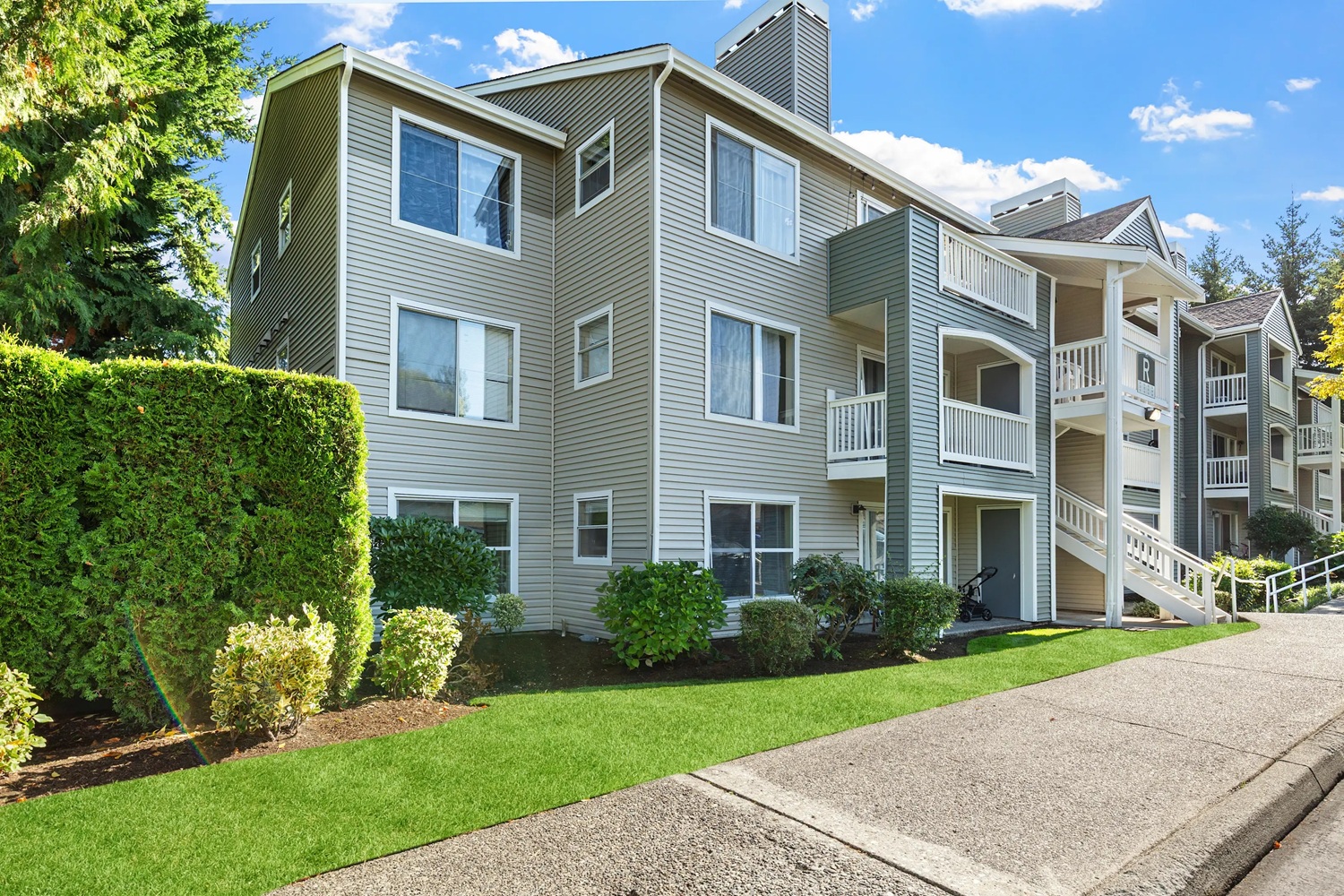 Gray apartment building with balconies, walkways, and lush green landscaping.