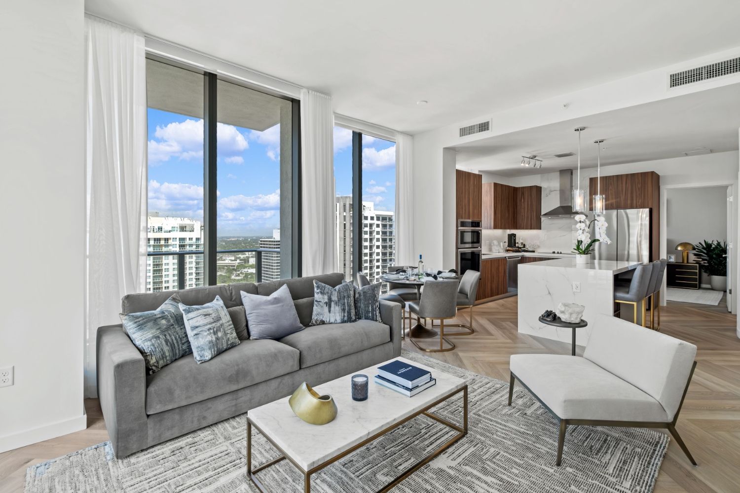 Modern living room with a gray sofa, chair, and large windows showcasing a city view.