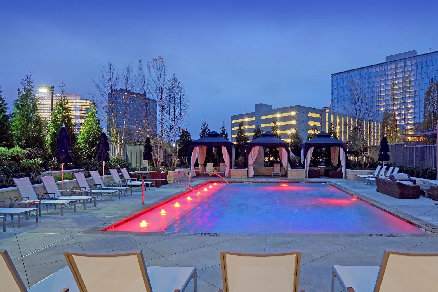 Outdoor pool area at dusk with illuminated water and lounge chairs.