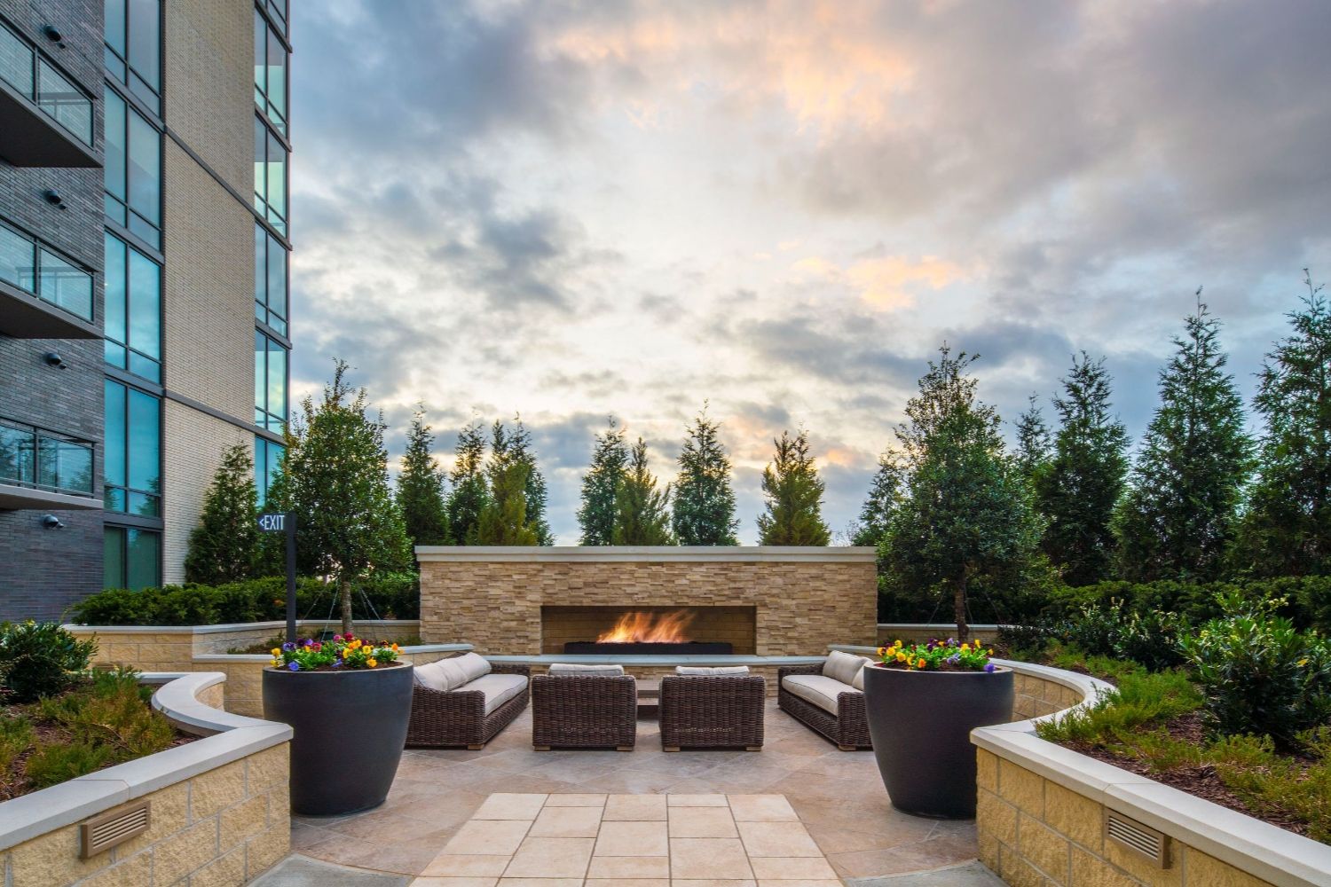 Outdoor seating area with a fire pit, surrounded by plants and a cloudy sky at sunset.