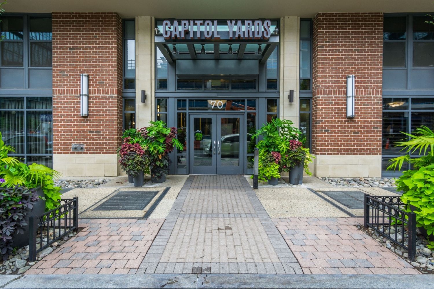 Entrance of a modern building with glass doors, brick walls, and surrounding greenery.