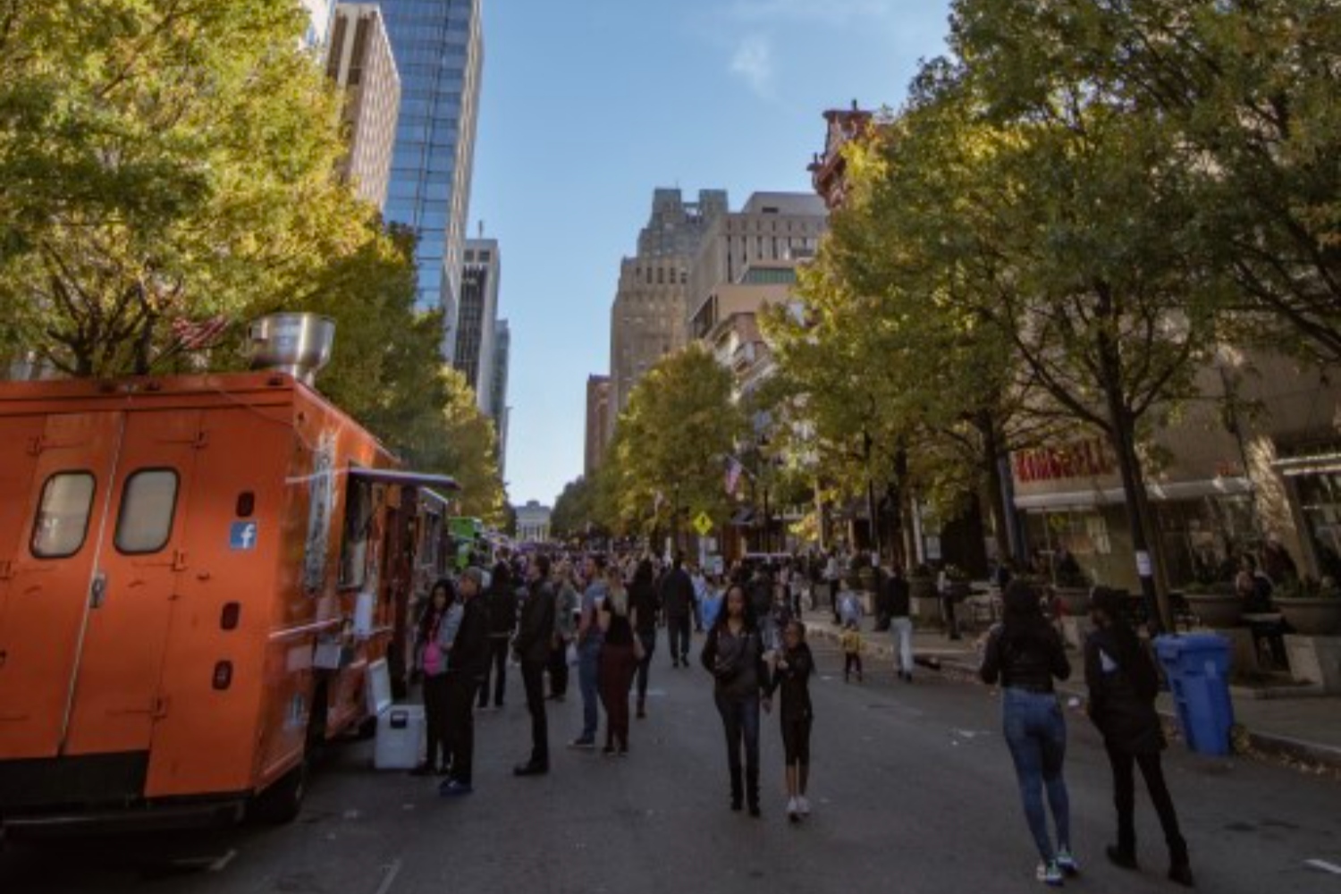 Busy street scene with a food truck and people walking among trees and buildings.
