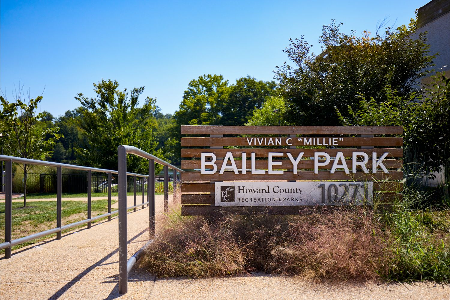 Bailey Park entrance sign beside a paved path, metal railing, and landscaped greenery.