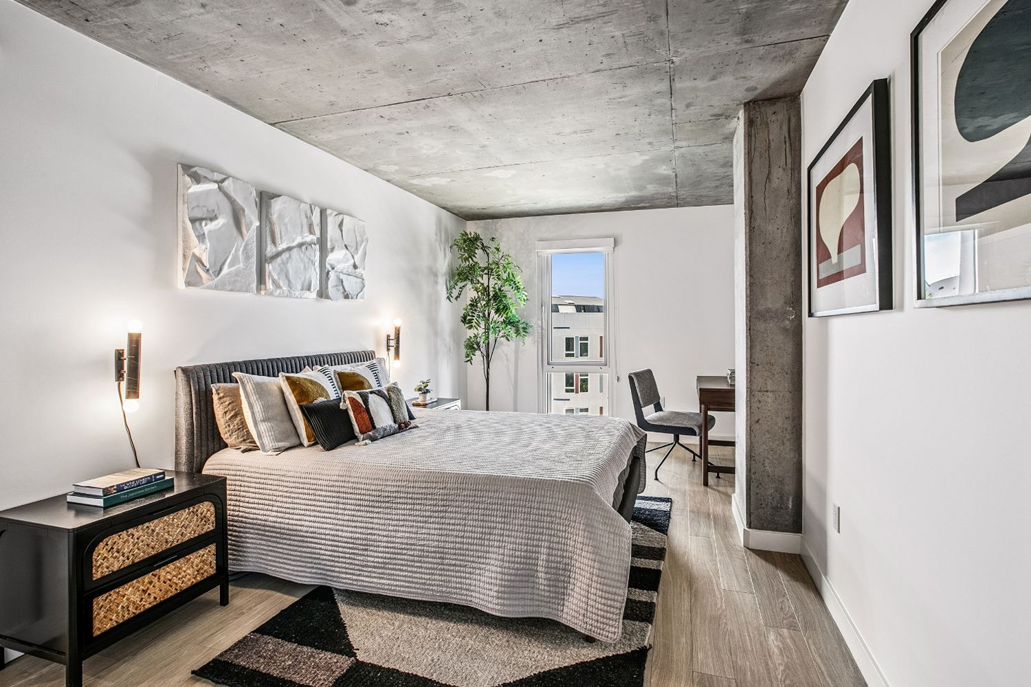 Bright bedroom at The Brady with an exposed concrete ceiling, wood-style flooring, large window, upholstered bed, and modern furnishings.