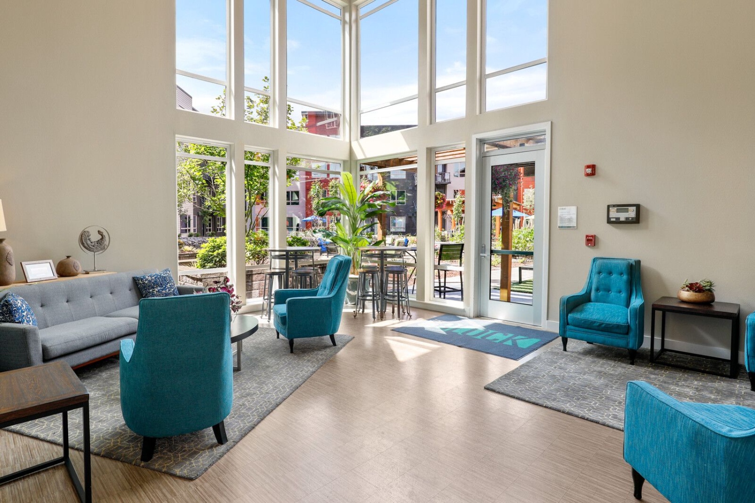 Bright lounge with teal chairs, a sofa, and large windows showing outdoor greenery.