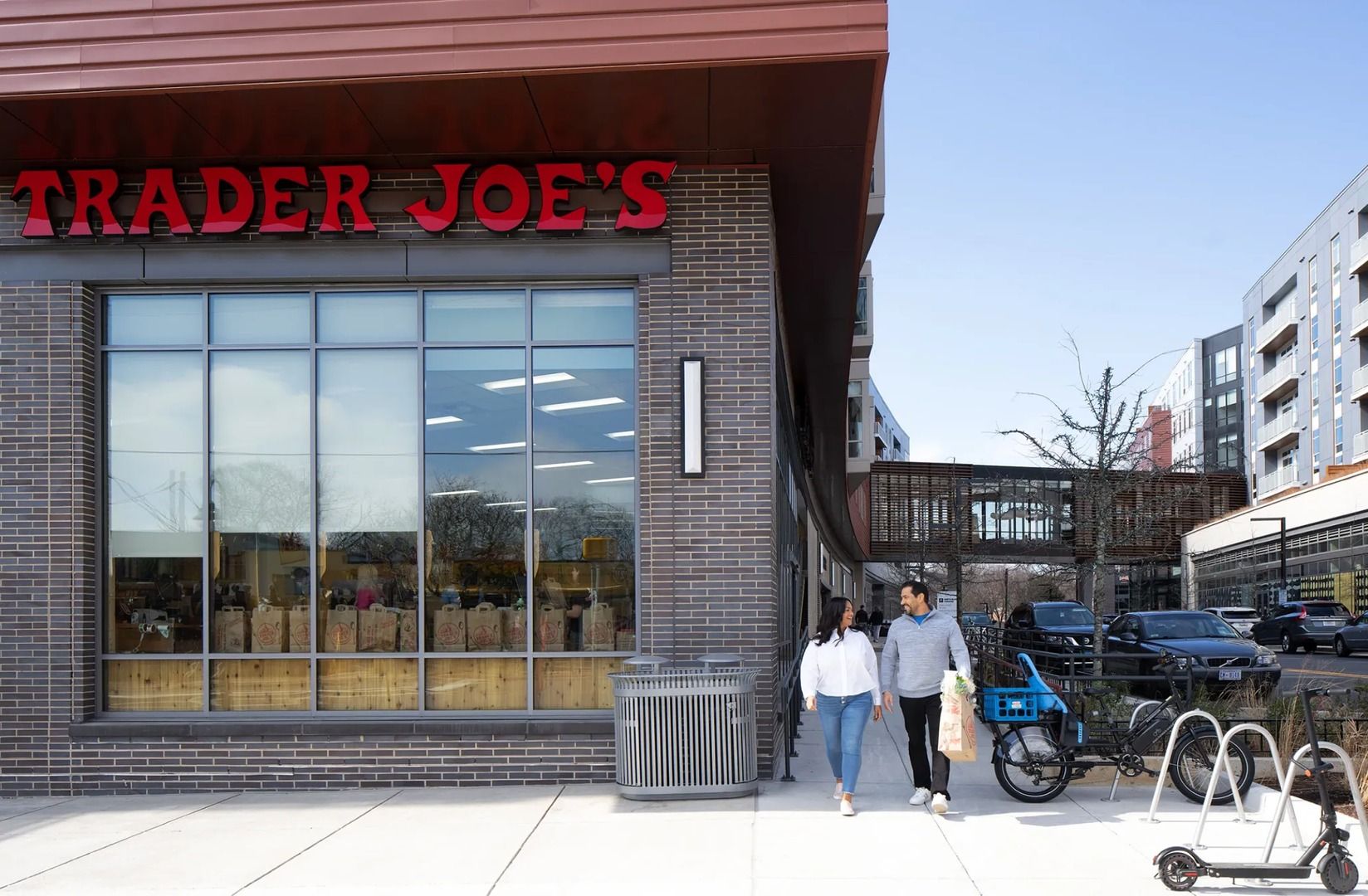 Two people walk past a Trader Joe's store with large windows and a modern exterior.