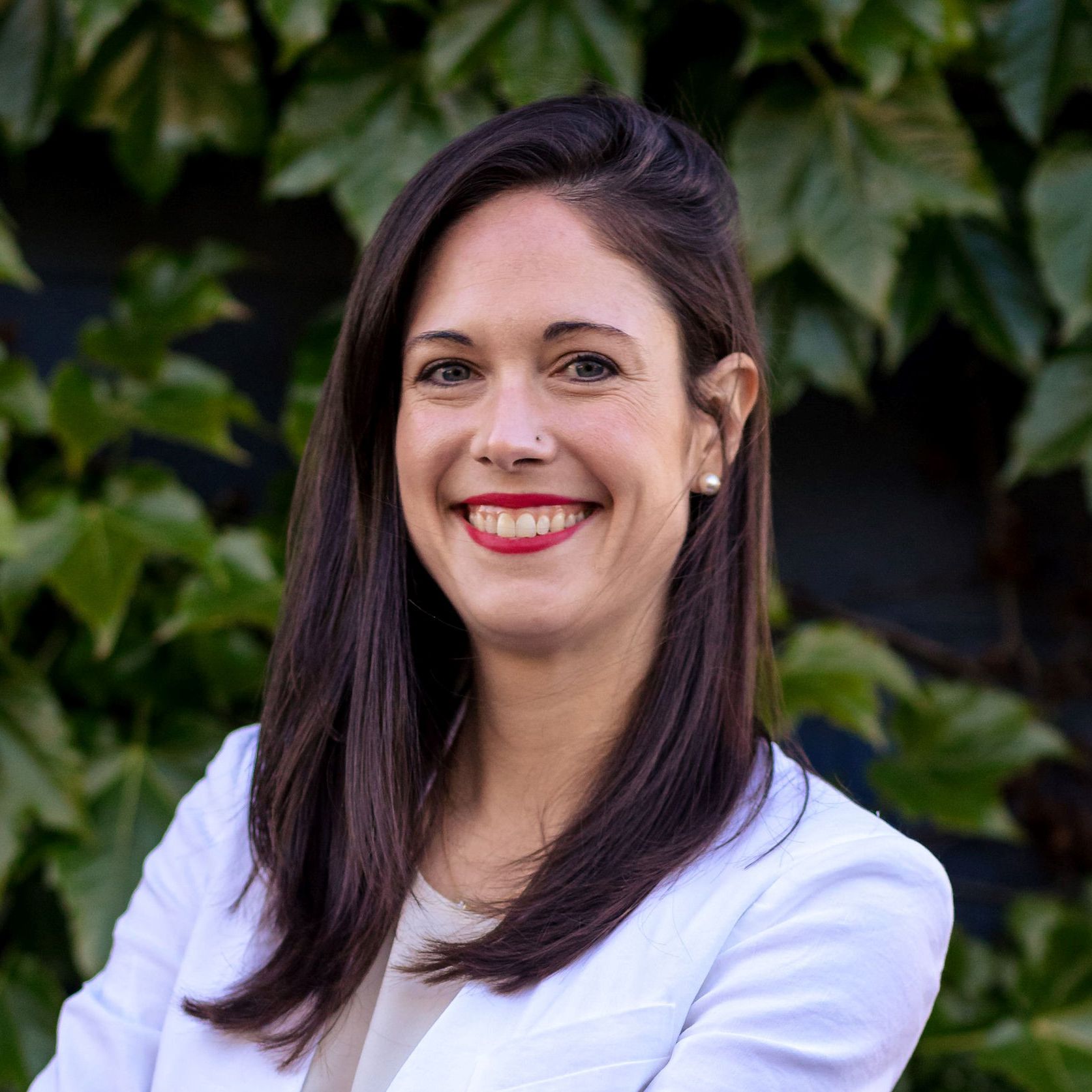 Smiling woman in a white blazer standing against a backdrop of green foliage.