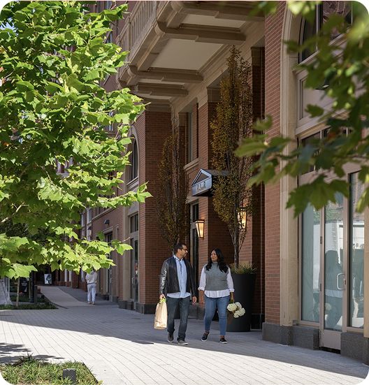 Two people walk along a sidewalk by storefronts and trees.