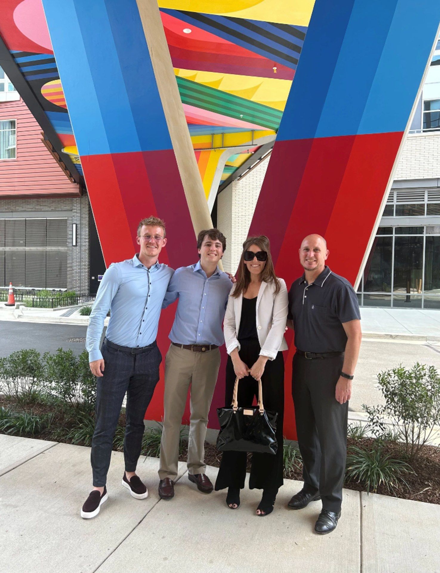 Four people pose together under a colorful architectural structure outdoors.