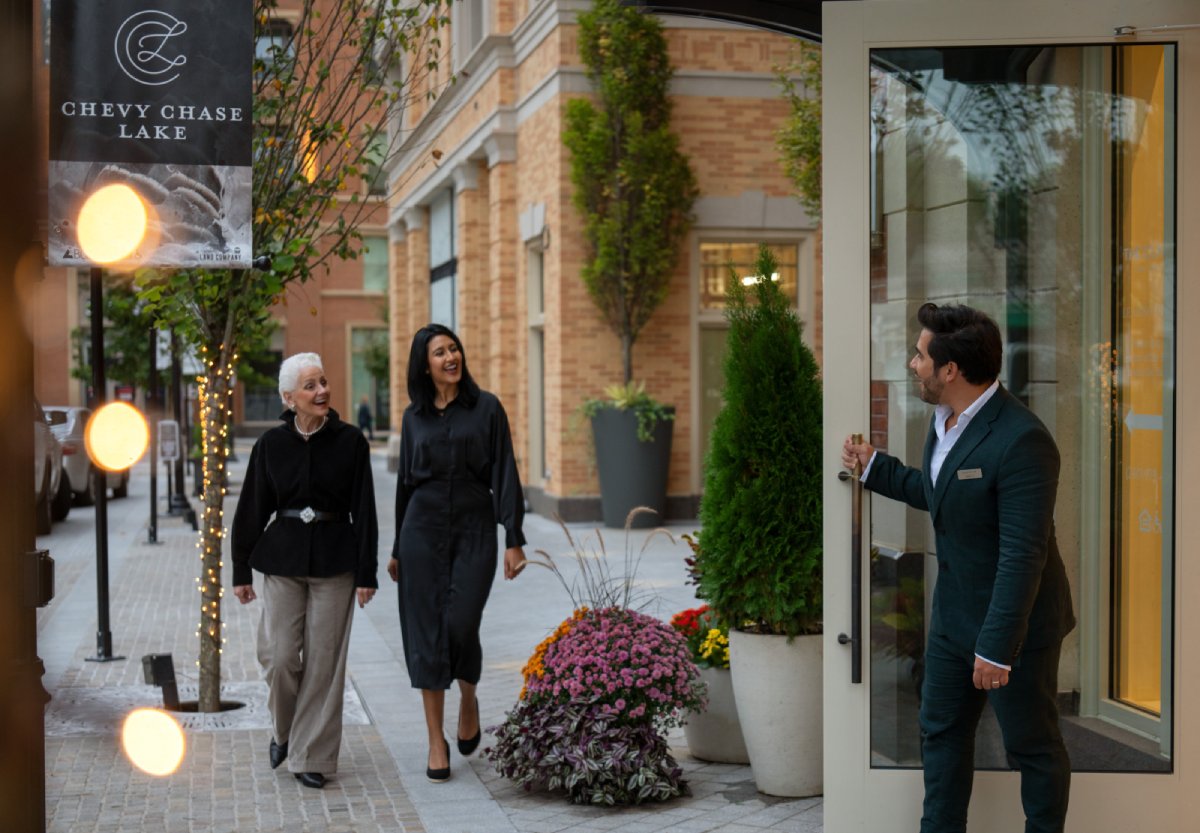 Two women walk past a doorway where a man is greeting them at a stylish entrance.