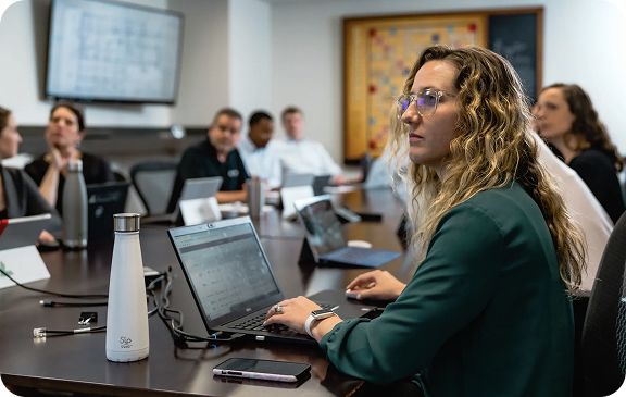 A woman with glasses focuses on a laptop in a meeting with several participants.