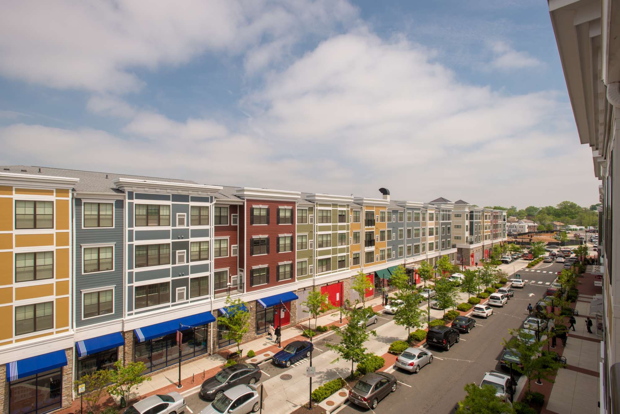 Modern multi-story buildings line a sunny street with parked cars and green trees.