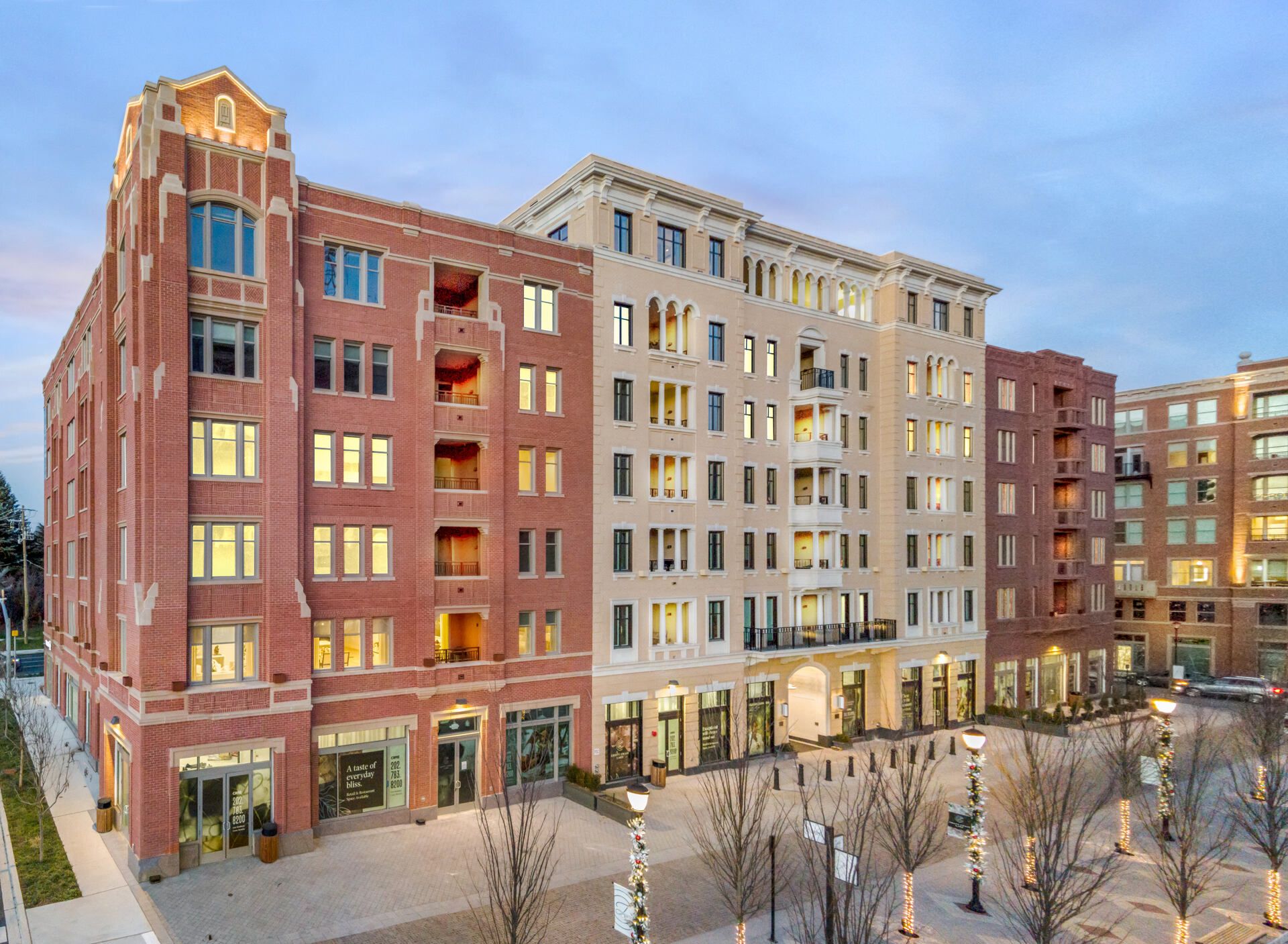 Modern multi-story building with a mix of brick and stucco exteriors, lit windows, and trees.